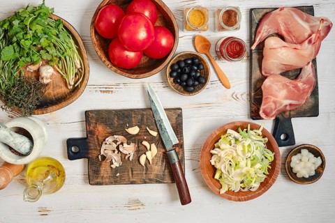 Fresh tomatoes, herbs, and spices are laid out on wooden boards in a kitchen. A knife and cutting