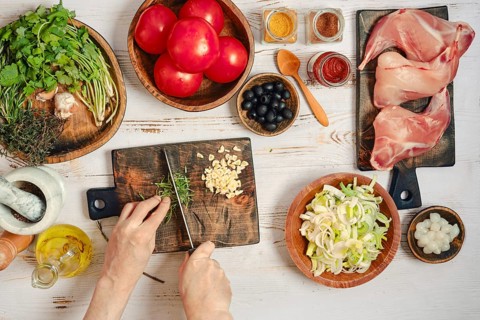 Hands are cutting rosemary on a wooden board. Fresh tomatoes, herbs, spices, and rabbit legs are