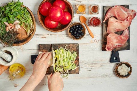 Fresh vegetables, meats, and spices are arranged on a table. Someone is chopping onions with a