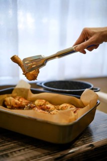 A hand lifts a piece of grilled chicken drumstick from a tray. The scene shows a cozy kitchen setup