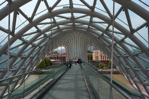 View through the modern glass bridge across a river with urban surroundings, Peace Bridge, Tbilisi,
