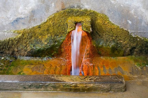 Rust-brown water flows from a spring in a concrete wall surrounded by eroded mineral deposits,