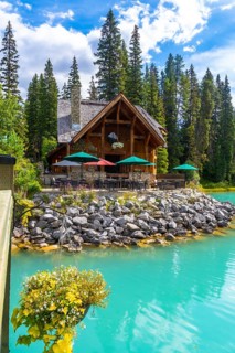 Wooden lodge with patio furniture and umbrellas reflecting in turquoise water on the shore of