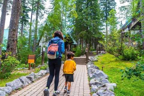 Tourists walking on a paved path approaching emerald lake lodge, surrounded by lush forest and