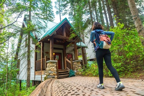 Female tourist with backpack walking on a paved path. Admiring a wooden cabin in the forest near