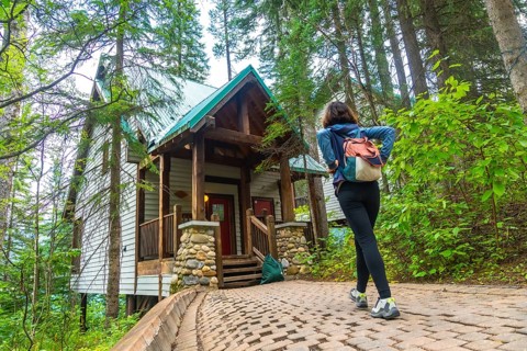 Female tourist with backpack walking up cobblestone path to a wooden cabin surrounded by lush green