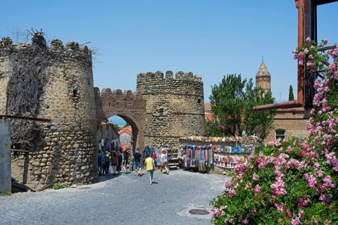 An old city gate with fortress wall and tower, animated by tourists, flanked by colorful stalls and