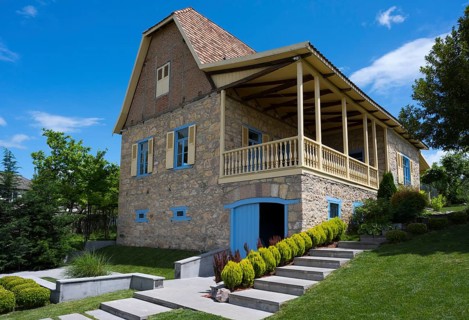 Traditional stone house with veranda and shutters, surrounded by well-kept garden under blue sky,