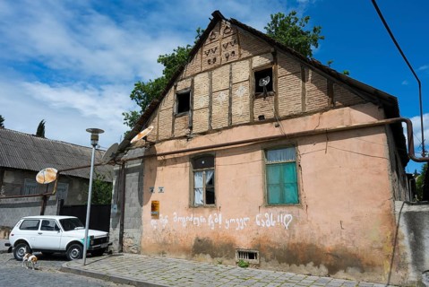 A dilapidated house with leaning antennas and graffiti, next to a parked car under cloudy sky,