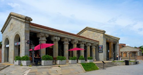 Historic stone building with ancient columns and red umbrellas under cloudy sky, Mtskheta,