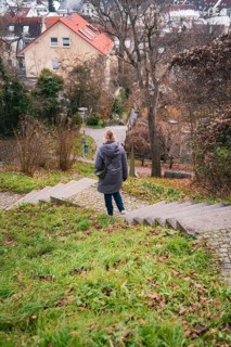 A woman in a cloak walks down an overgrown staircase surrounded by trees and houses, Herrenberg,