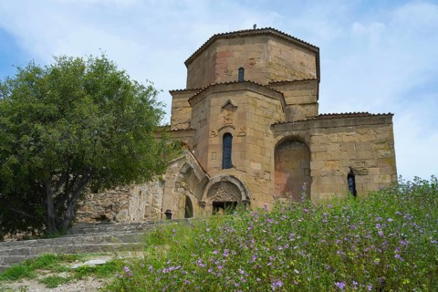 Stone church with decorated details surrounded by flowering plants and a large tree under a blue