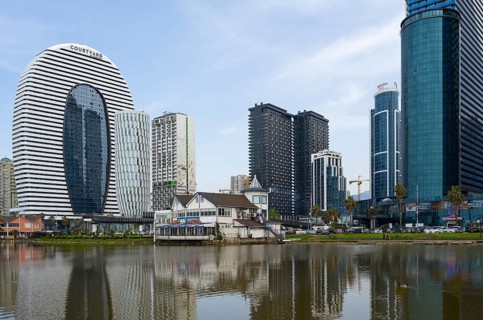 Modern high-rise buildings with glass facades are reflected in a calm lake under a cloudy sky,