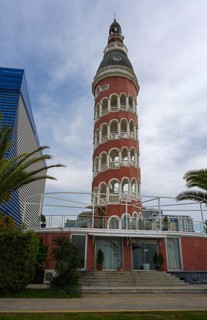 A historic brick tower with round arches stands next to palm trees and modern architecture under a