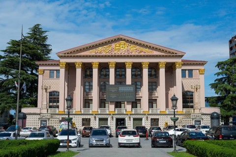 An imposing building with ancient columns and golden decoration, cars parked in front of it, Ilia