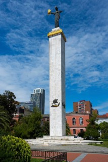 High column with statue in front of modern buildings under clear sky, statue of Medea, Europe