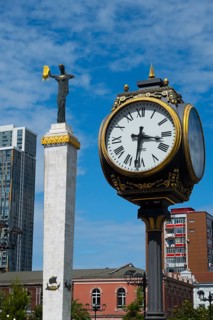 Large clock and statue in front of modern buildings under bright blue sky, clock and statue of