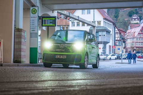 Green shuttle at a covered stop, surrounded by half-timbered houses, Deer E-Mobility Shuttle