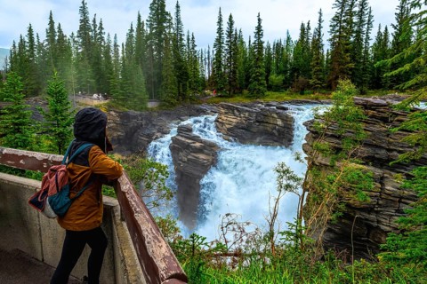 Tourist in a hooded jacket and backpack leaning on a wooden railing, admiring the powerful
