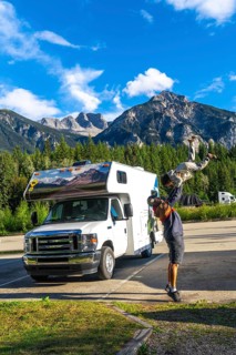 Father playfully lifting his son in the air in front of their camper van, enjoying a scenic stop