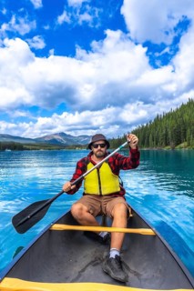 Tourist wearing sunglasses and a life jacket paddling a canoe on turquoise maligne lake under a