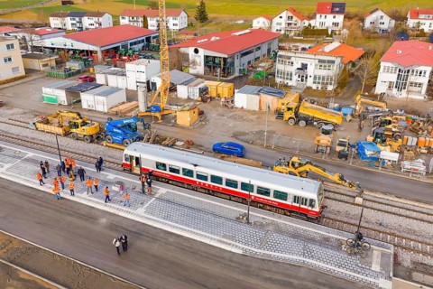 Workers work next to a train and tracks at a construction site with adjacent buildings, historic