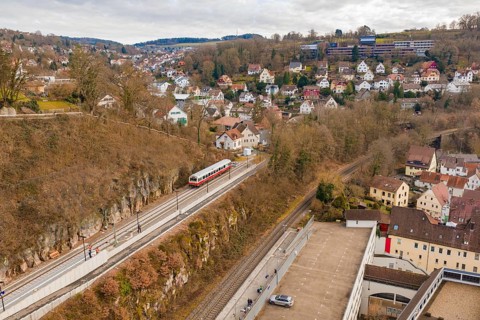 A train travels through a hilly area with urban houses and winter trees under a cloudy sky,