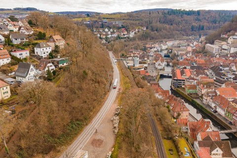 A train moves through a picturesque town, which is located along a hill and near a riverbank,