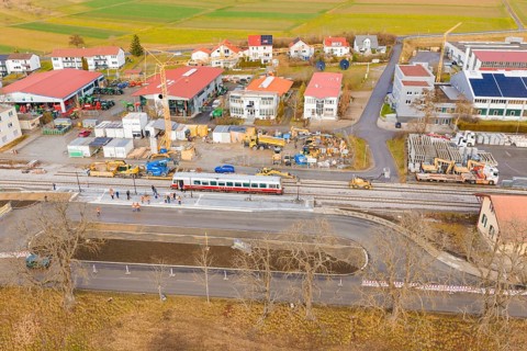 A construction site next to a train, surrounded by buildings and fields, is viewed from the air,