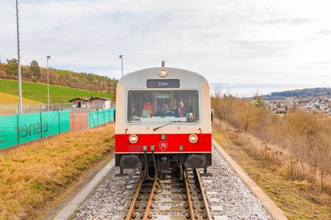 Train with passengers and the inscription 'Calw' on the rails, with landscape in the background,