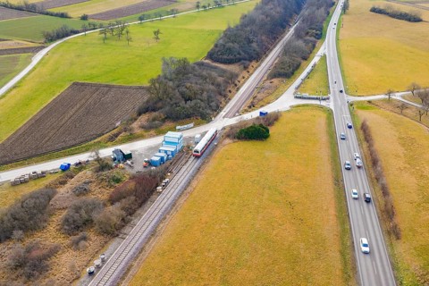 Train travels near a country road in a rural area with fields and vehicles, historic event, first