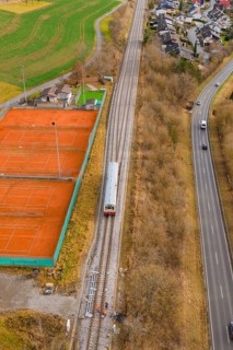 A train travels along railroad tracks past tennis courts, next to a busy road, historic event,