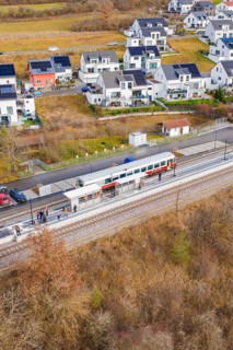 Train stops at a small train station next to modern houses surrounded by fields, historic event,