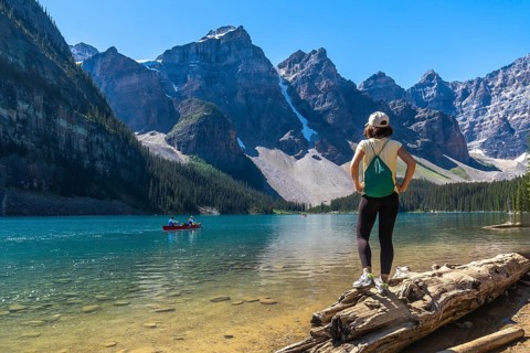 Female tourist standing on a log, admiring turquoise waters of moraine lake with canoers,