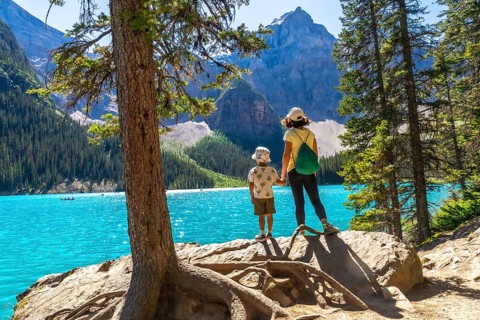 Mother and son holding hands on a rock by a tree, admiring the turquoise waters of moraine lake,