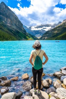 Young woman backpacker standing on rocks admiring stunning turquoise lake and surrounding mountains