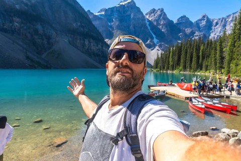 Backpacker taking a selfie with turquoise lake and canoes with the canadian rockies in the