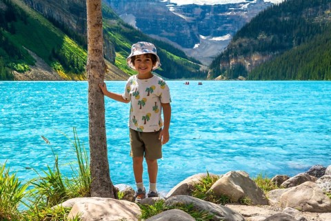 Young child standing by a tree on the rocky shore of turquoise lake louise, taking in the