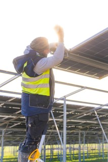 Worker mounts solar panel on scaffolding, stands on ladder with yellow safety vest, energy