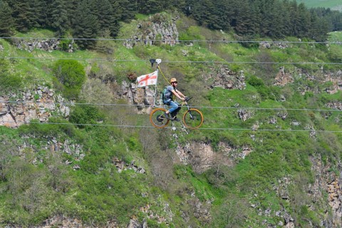 Person riding a bicycle on a rope in front of a wooded rocky landscape in the mountains, Dashbashi