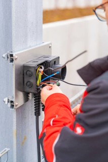 Person wearing a red jacket assembles electrical cables at a metal station, parking garage