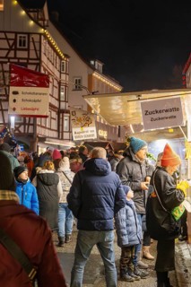 Night market visitors at an illuminated cotton candy stand in front of half-timbered houses,