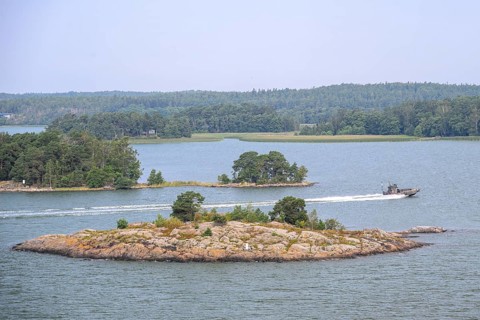 Small, wooded island in calm water with passing patrol boat boat, marine speedboat, Aaland Islands,