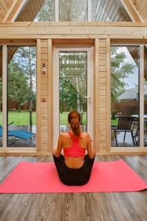 Back view of a woman sitting cross-legged on a pink yoga mat inside a wooden cabin. Outside, there