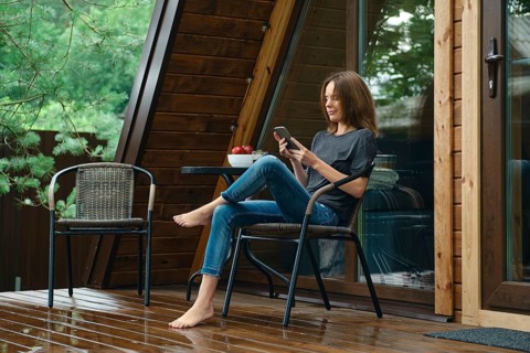 Barefoot woman sits on a chair at a table on a wet wooden porch and looks at a mobile phone. The