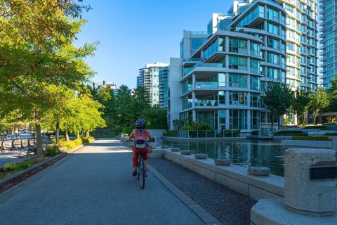 Woman wearing a helmet and backpack cycling on a paved path in an urban park surrounded by green