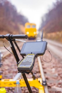 A technical device with a screen in front of a yellow vehicle on the rails, tamping machine on