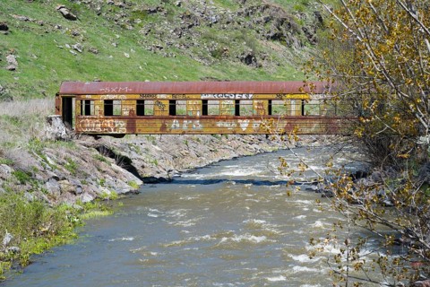 A rusted train car with graffiti stands over a flowing river in a green, hilly landscape, discarded
