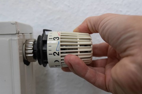 A hand grabs the thermostat head of a radiator to adjust the temperature. This image symbolizes