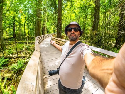 Photographer with hat and sunglasses taking a selfie while walking down a wooden walkway in a lush